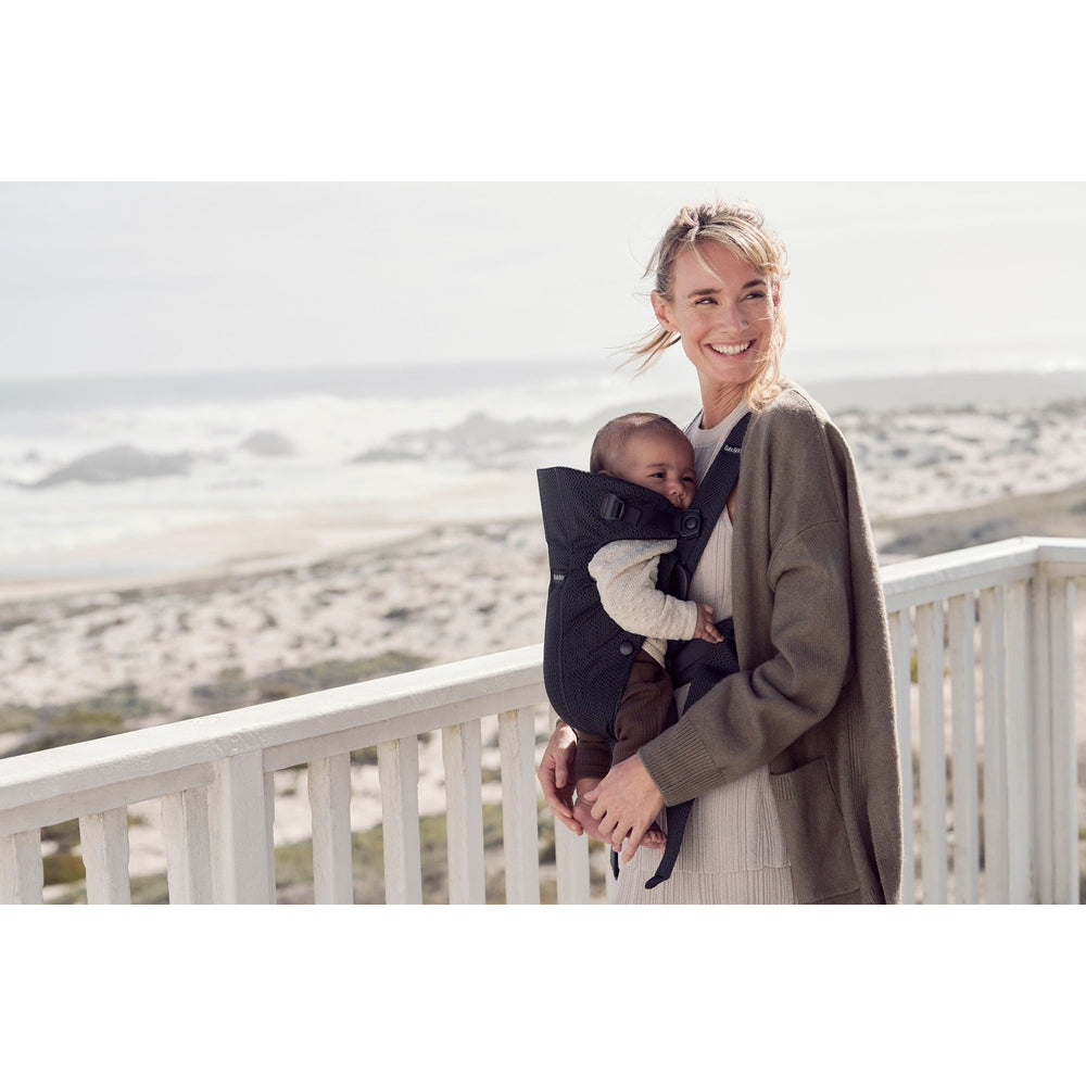 BabyBjörn Baby Carrier Mini worn by a smiling mom on a seaside boardwalk, holding a baby in the dark gray carrier.