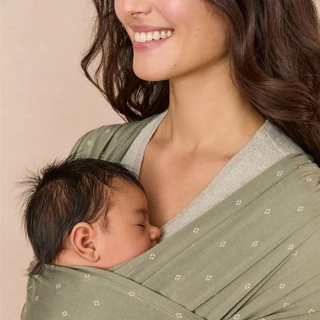 Ergobaby Aura Sustainable Knit baby wrap in sage green, worn by smiling mother with sleeping infant.