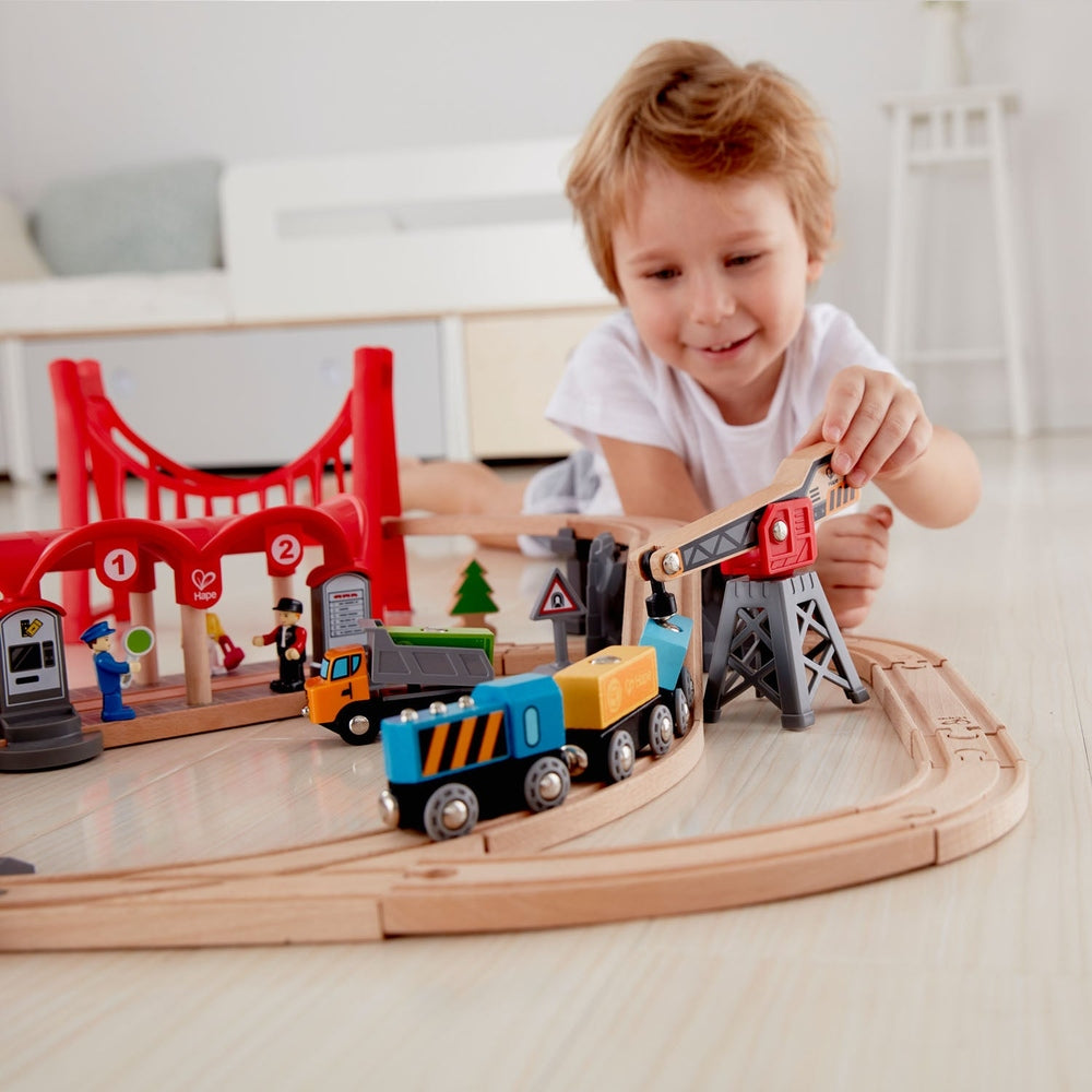 Hape Busy City Rail Set; child builds on curved wooden tracks with a crane and colorful trains.