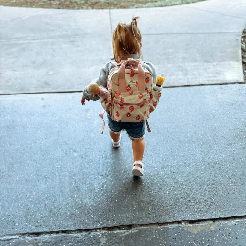 Itzy Ritzy backpack strawberry print with pink straps worn by a toddler walking outdoors with a stuffed toy.