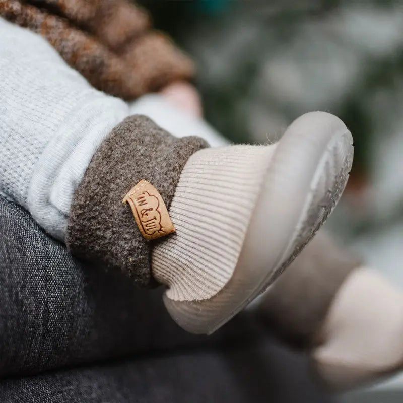 Jan & Jul Cozy Sock Shoes Oat, baby’s foot snug in soft beige knit sock with cushioned sole and leather tag.