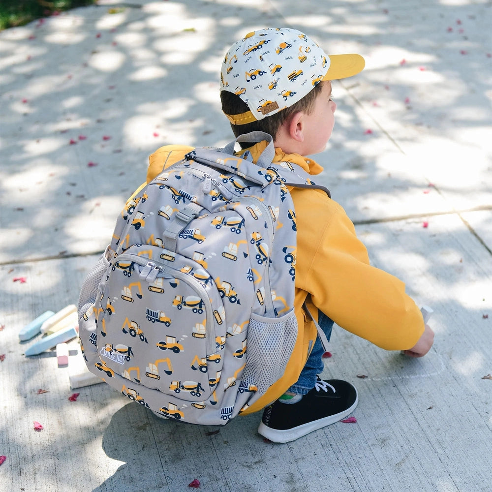Jan & Jul Kids Backpack in light gray with yellow construction truck print, worn by a child crouching on the sidewalk reaching for chalk.