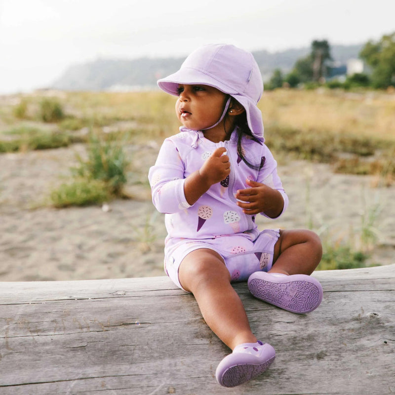 Jan & Jul Kids One Piece UV Sun Suit Lavender Ice Cream with matching hat; toddler sits on a wooden railing by the beach.