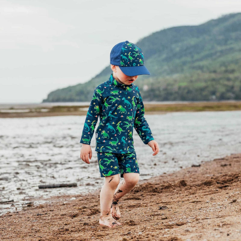 Jan & Jul Kids UV Rashguard Set Dinoland: blue dinosaur-print long-sleeve top with matching shorts, toddler walking barefoot on the beach.