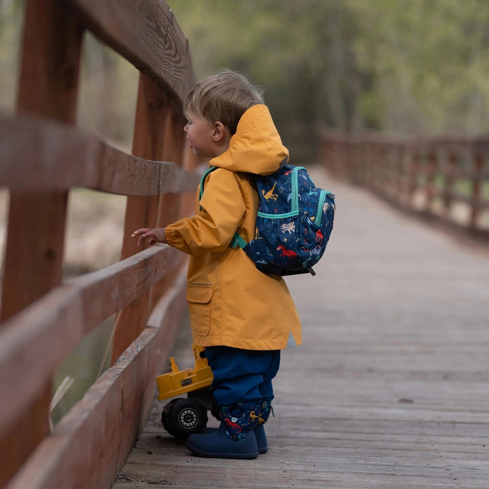 Jan & Jul Kids Mini Backpack in blue dinosaur print worn by a toddler in a yellow raincoat on a wooden bridge.
