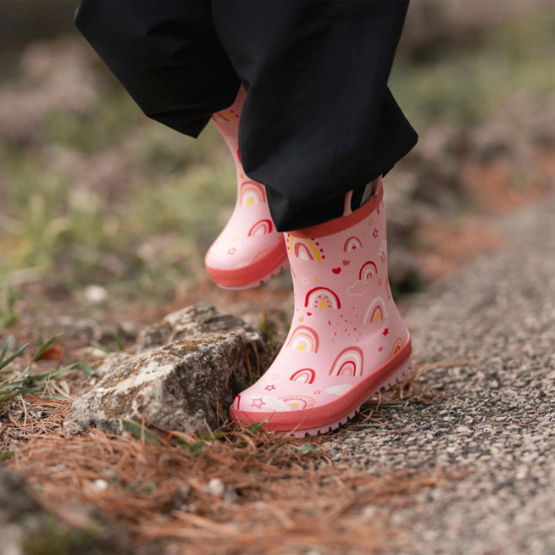 Jan & Jul Puddle Dry Rain Boots Pink Rainbow worn by a toddler, stepping over a rock on a gravel path.