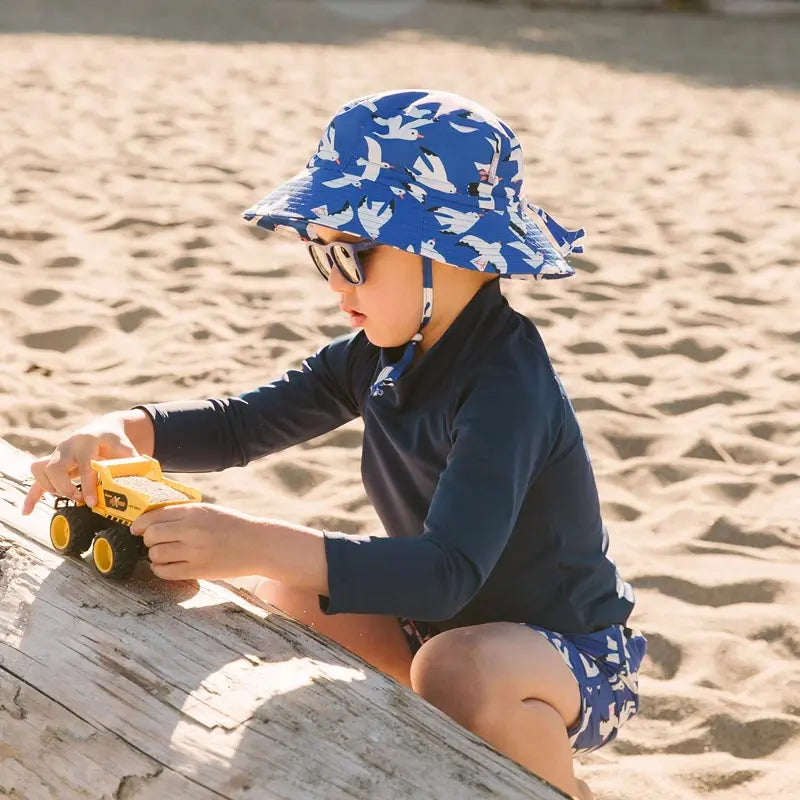 JAN & JUL water-repellent blue seagull bucket hat worn by a toddler at the beach, playing with a yellow toy truck.