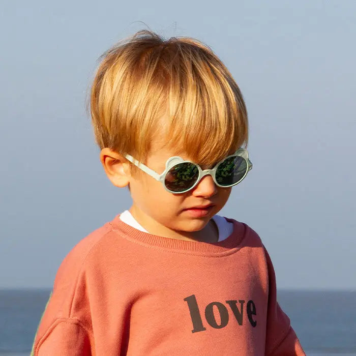 Ki Et La Sunglasses Ourson Almond Green worn by a child at the beach, round white frames, dark lenses.