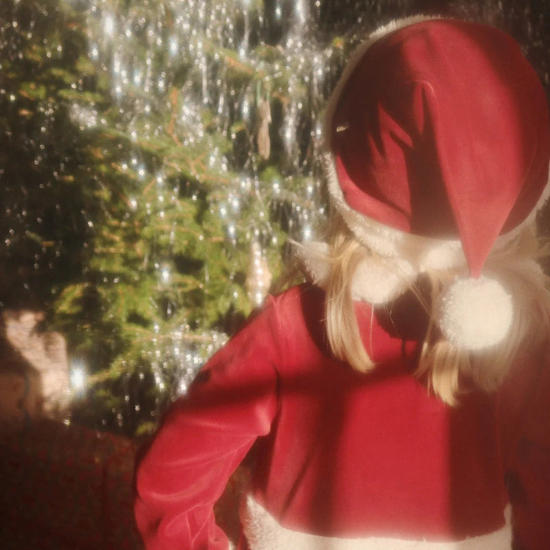 Konges Slojd Christmas Hat Red Dahlia on a toddler facing away in a red coat beside a Christmas tree.
