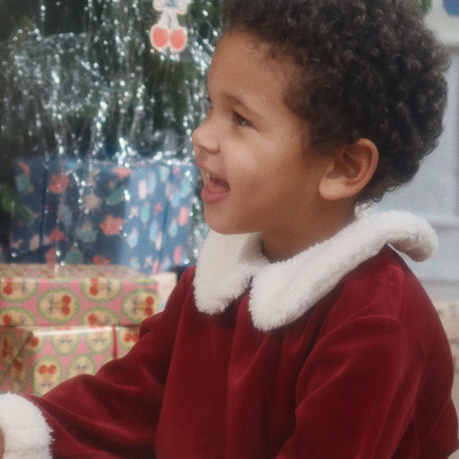 Konges Slojd Christmas Set Red Dahlia toddler outfit in red velvet with white faux fur trim, worn by a smiling child near wrapped gifts.