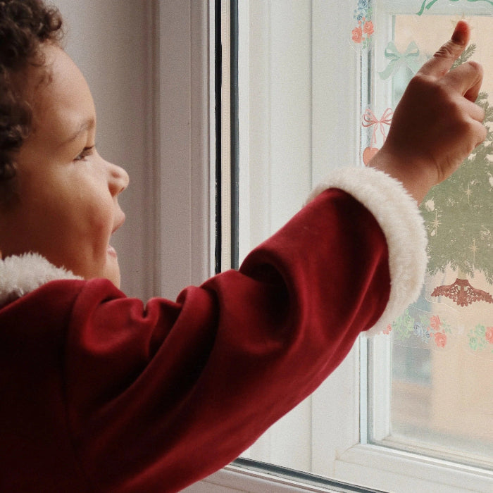 Konges Slojd Christmas Set Red Dahlia: toddler in red velvet with white fur cuffs reaches for festive window decals.