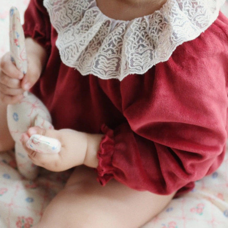 Konges Slojd Venola Romper Red Dahlia worn by a baby in burgundy fabric with white lace collar, gripping a soft toy.