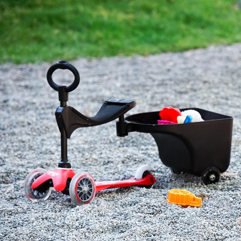 Micro Balance Bike Trailer - red balance bike hitched to a black trailer on gravel with toys inside.