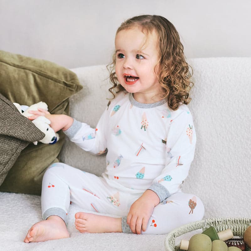 Nest Designs Bamboo Jersey Two-Piece Long Sleeve Pajama Set Ice Cream Summer; toddler in white pastel-print PJs sits on sofa playing with a plush toy.