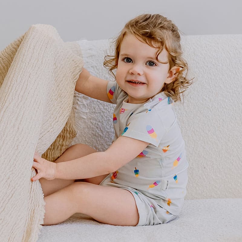 Nest Designs Bamboo Jersey Two-Piece Short Sleeve Pj Set Summer Pops: toddler sits on a sofa in light gray PJs with colorful pops, grabbing a cream blanket