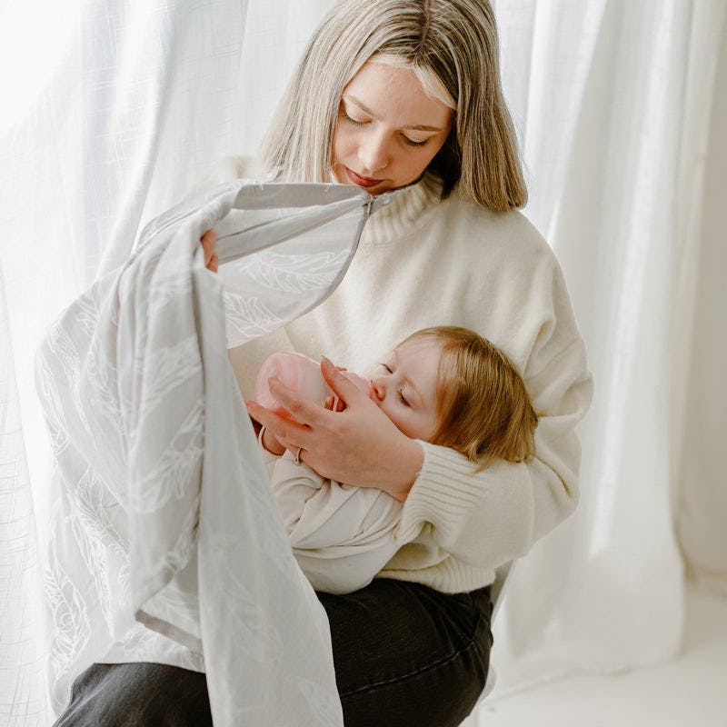 Nest Designs Bamboo Nursing Cover in beige drapes over mom while she nurses her baby.