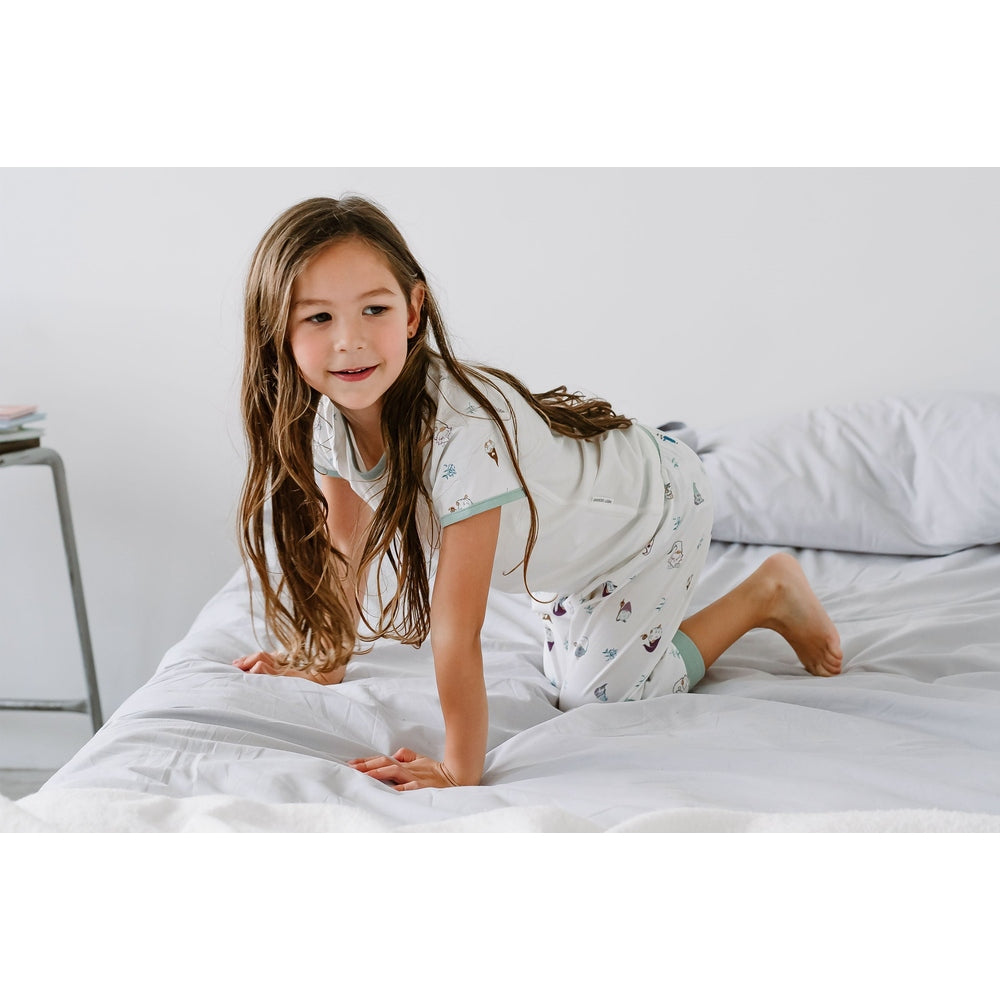 Nest Designs bamboo short-sleeve play set Oh Gnome, white pajamas with teal trim, girl on the bed smiling playfully.