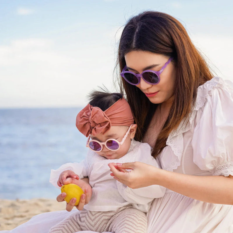 OLIVIO & CO Baby Round Sunglasses with coral blush frames; baby wearing purple-lens shades at the beach.