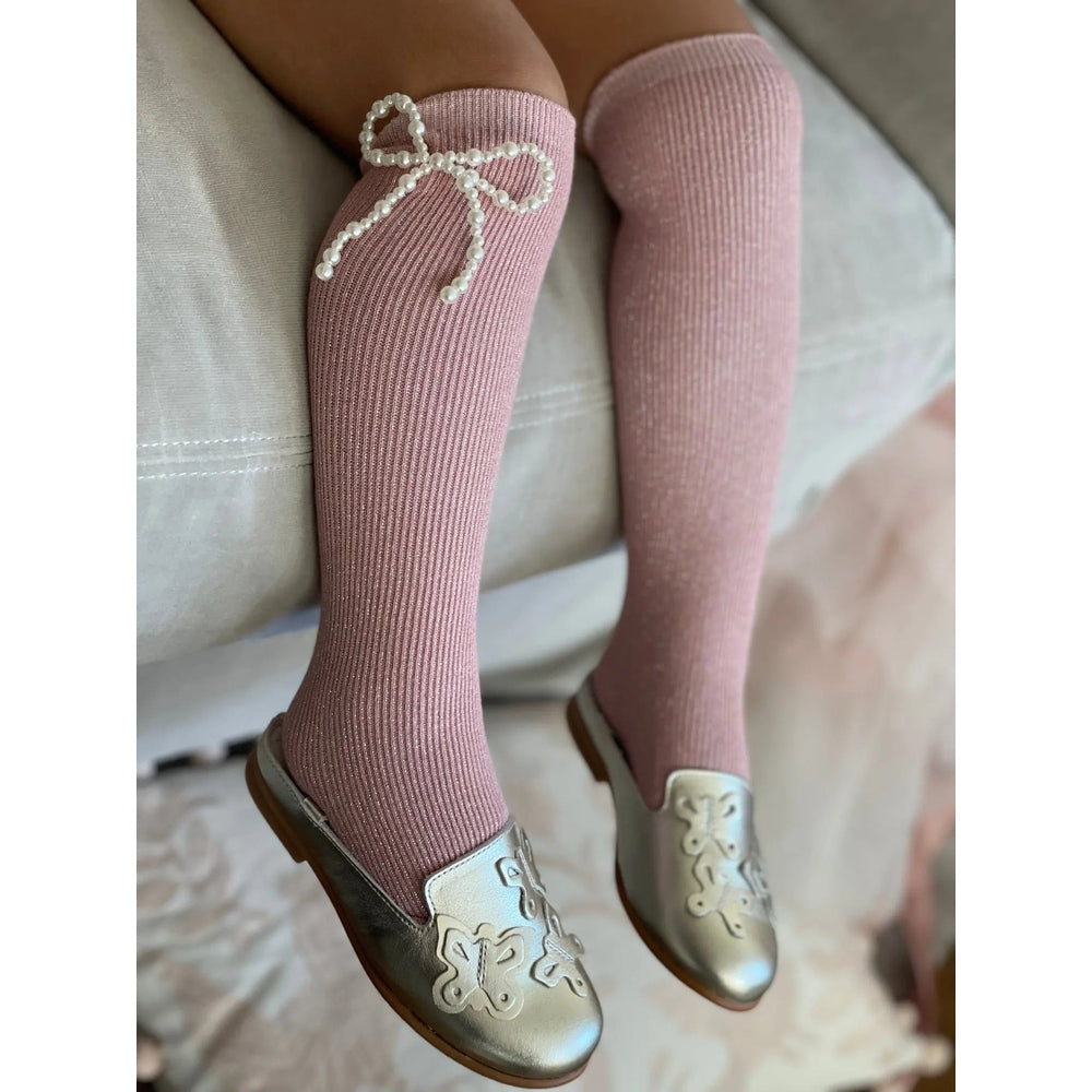 Petite Maison Kids Perla Shimmer Socks pink knee-highs with pearl bow detail, worn by a child sitting on a bed.