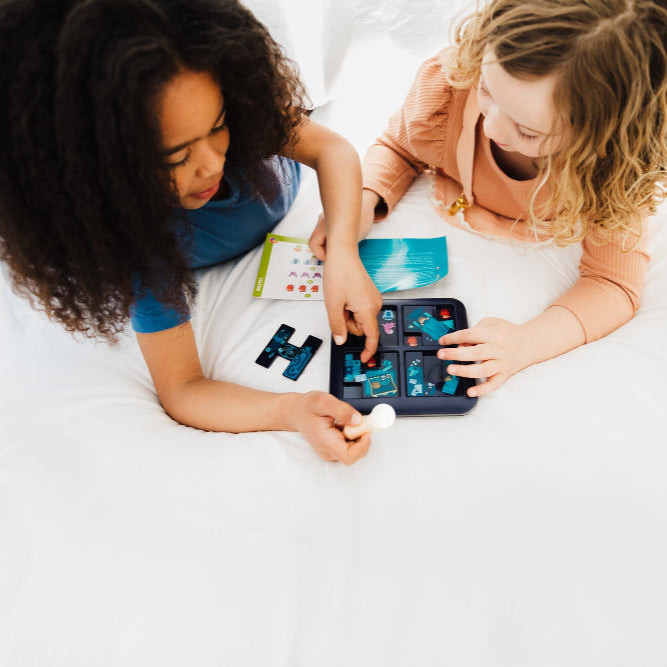 Smart Games Monsters Hide & Seek puzzle game; two kids on a bed playing with the handheld tile board.