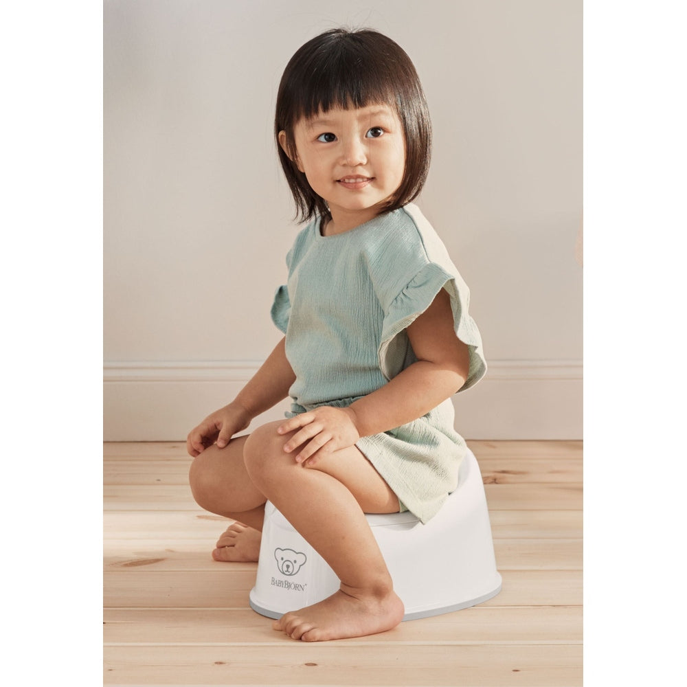 BabyBjörn Smart Potty, white with gray rim, toddler sits on it in a mint dress, demonstrating stability.