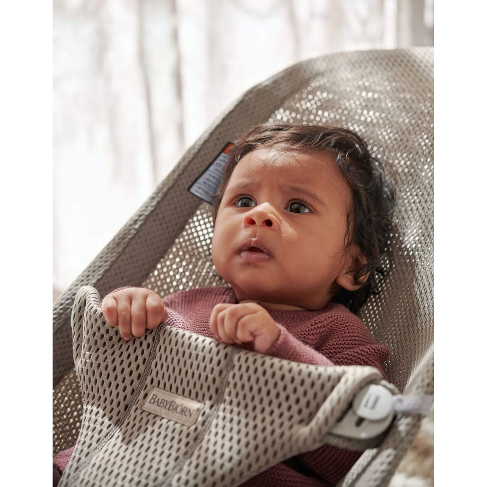 BabyBjörn Bouncer Bliss in grey mesh, infant seated and gripping the front edge in a curious moment.