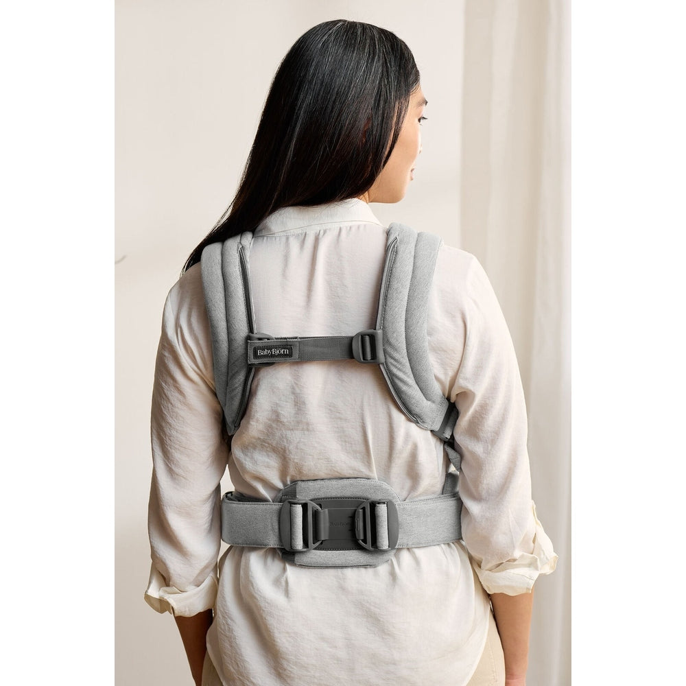 BabyBjörn Baby Carrier Harmony worn by a woman in a light shirt, featuring padded straps and a wide gray waist belt.