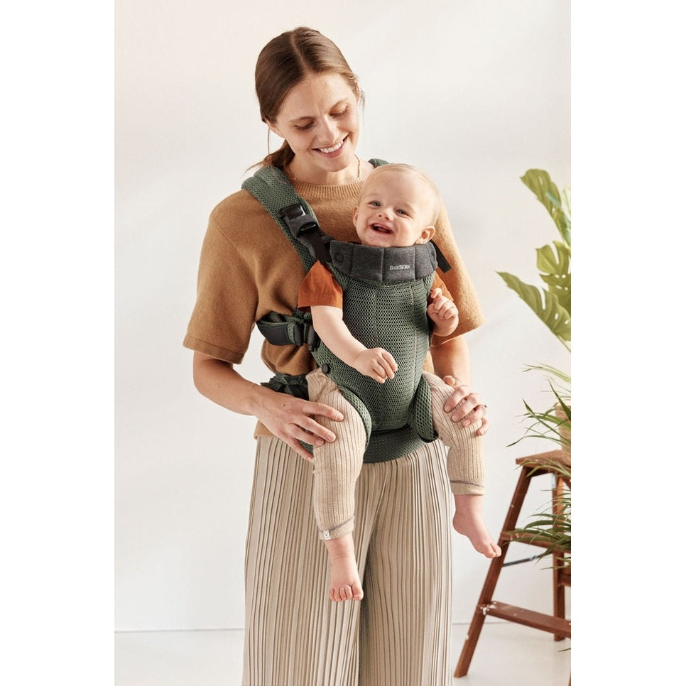 BabyBjörn Harmony baby carrier in green mesh worn by a smiling mom, showing secure waist belt and padded shoulder straps.