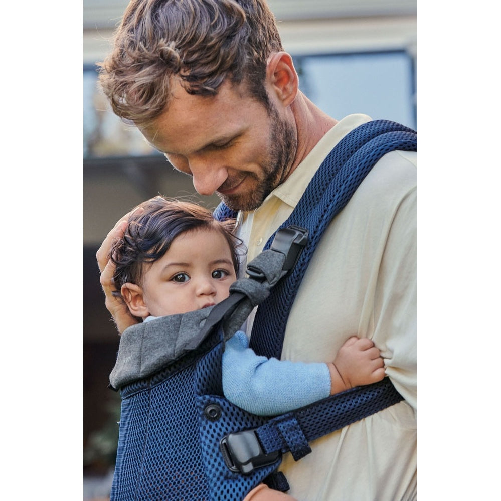 BabyBjörn Harmony baby carrier worn by a father in a beige shirt, baby facing inward and secured with a chest clip.