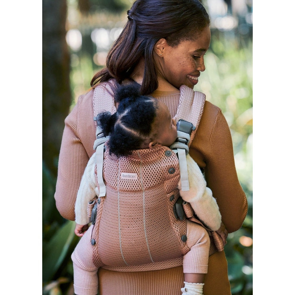 Mother outdoors wearing BABY BJÖRN Baby Carrier Harmony in blush pink, securely carrying infant on her back.