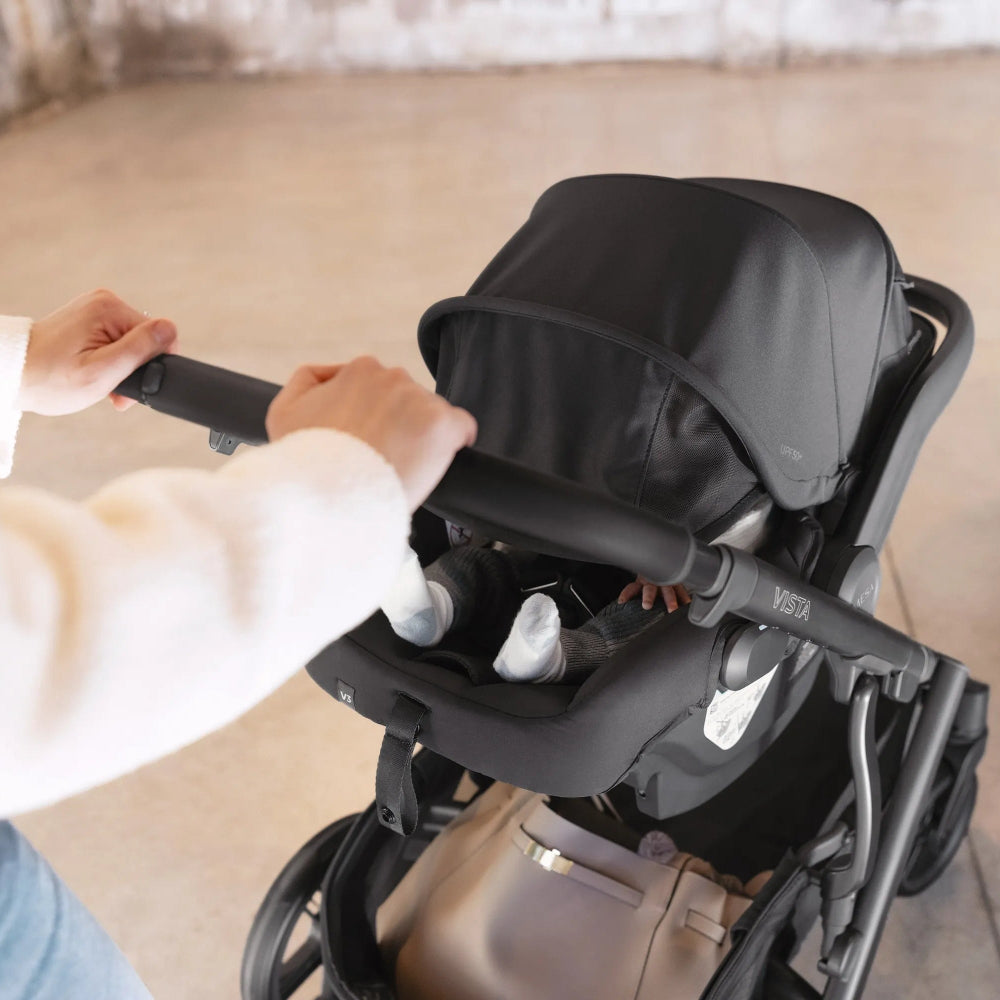 UPPAbaby Mesa V3 infant car seat secured to a black stroller, caregiver pushes the handlebar with both hands.