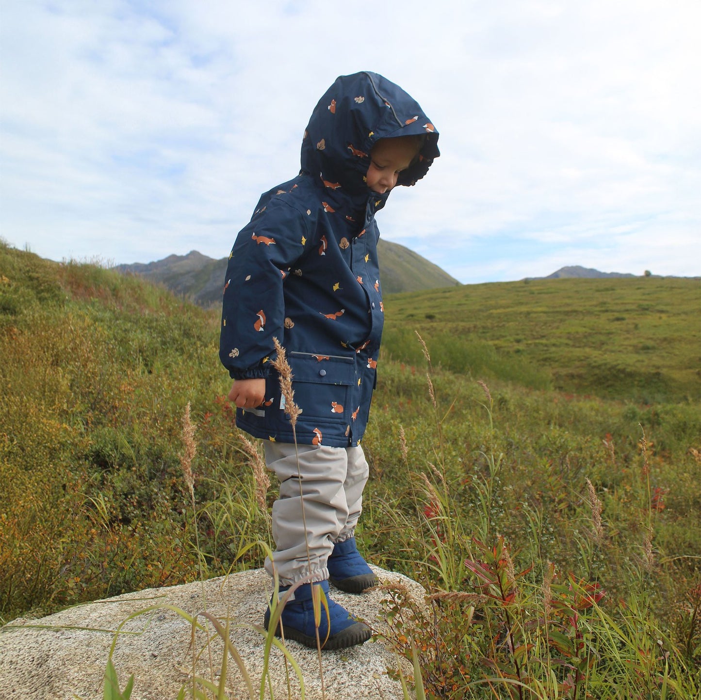 JAN & JUL Waterproof Cozy Dry Rain Jacket Navy Fox worn by a child with hood up, standing on a rock in a meadow.