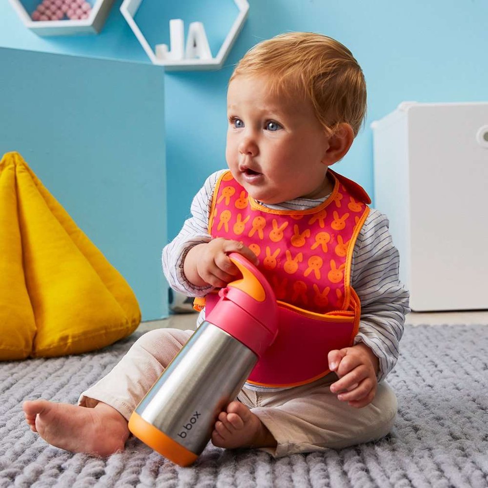 B.Box Insulated Bottle in pink cap and orange base, held by a toddler wearing an orange bib