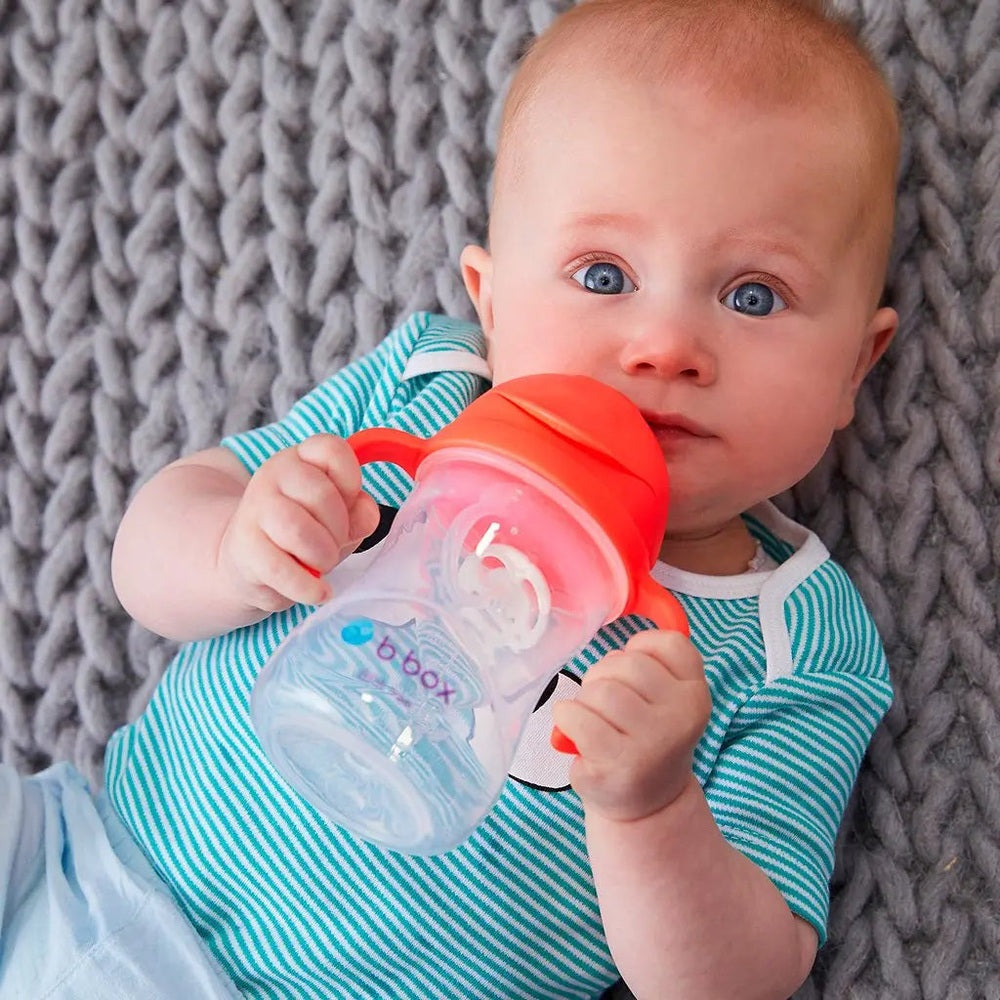 B.Box New Sippy Cup Matte Edition held by a baby in teal stripes, showing orange lid while drinking from a clear bottle.