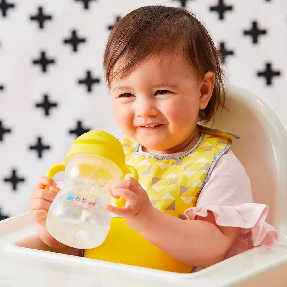 Baby smiles while gripping a B.Box New Sippy Cup Matte Edition, yellow handles on a clear bottle.