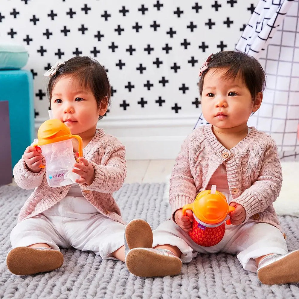B.Box New Sippy Cup Matte Edition; two toddlers in pink cardigans hold orange-lidded sippy cups while seated on a rug.