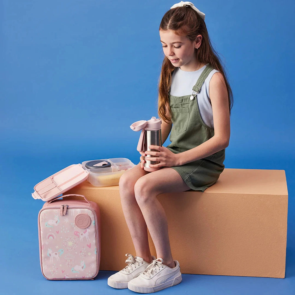 B.box Insulated Flexi Lunchbag pink unicorn pattern; girl sits on a block, holding a metal bottle with the lunch bag nearby.