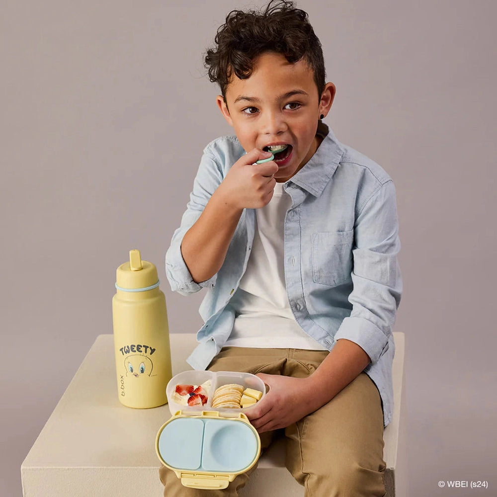 B.box Insulated Flip Top 1L water bottle in pastel yellow, with a child sipping from the flip-top spout beside a lunchbox.