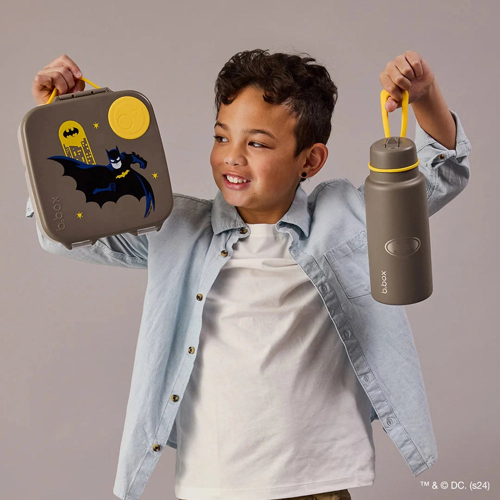 B.box Insulated Flip Top 1L water bottle in gray with yellow handle, held by a kid beside a Batman lunchbox