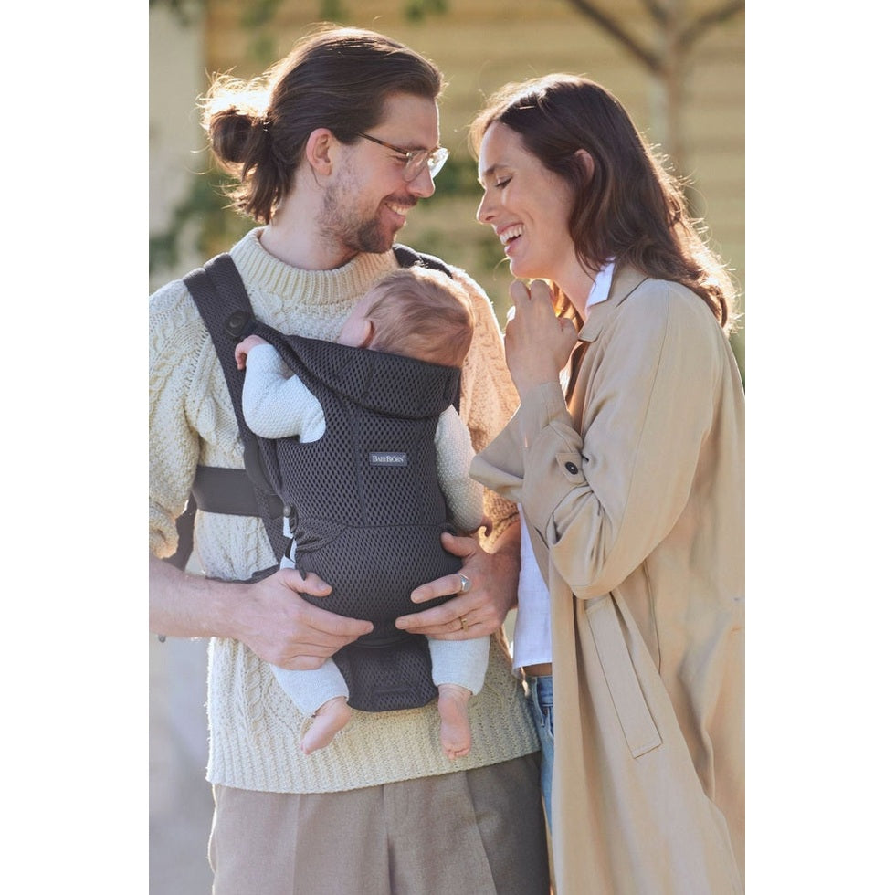 BabyBjörn Baby Carrier Free in dark-grey mesh worn by dad holding baby chest-to-chest while mom smiles outdoors.