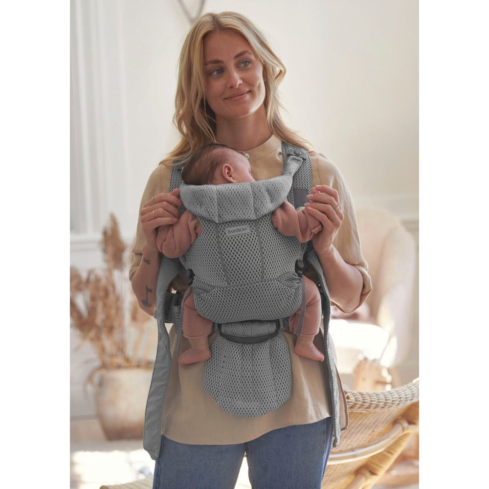 Mother wearing BABY BJÖRN Baby Carrier Free in gray mesh, securely holding a newborn in a cozy home setting.