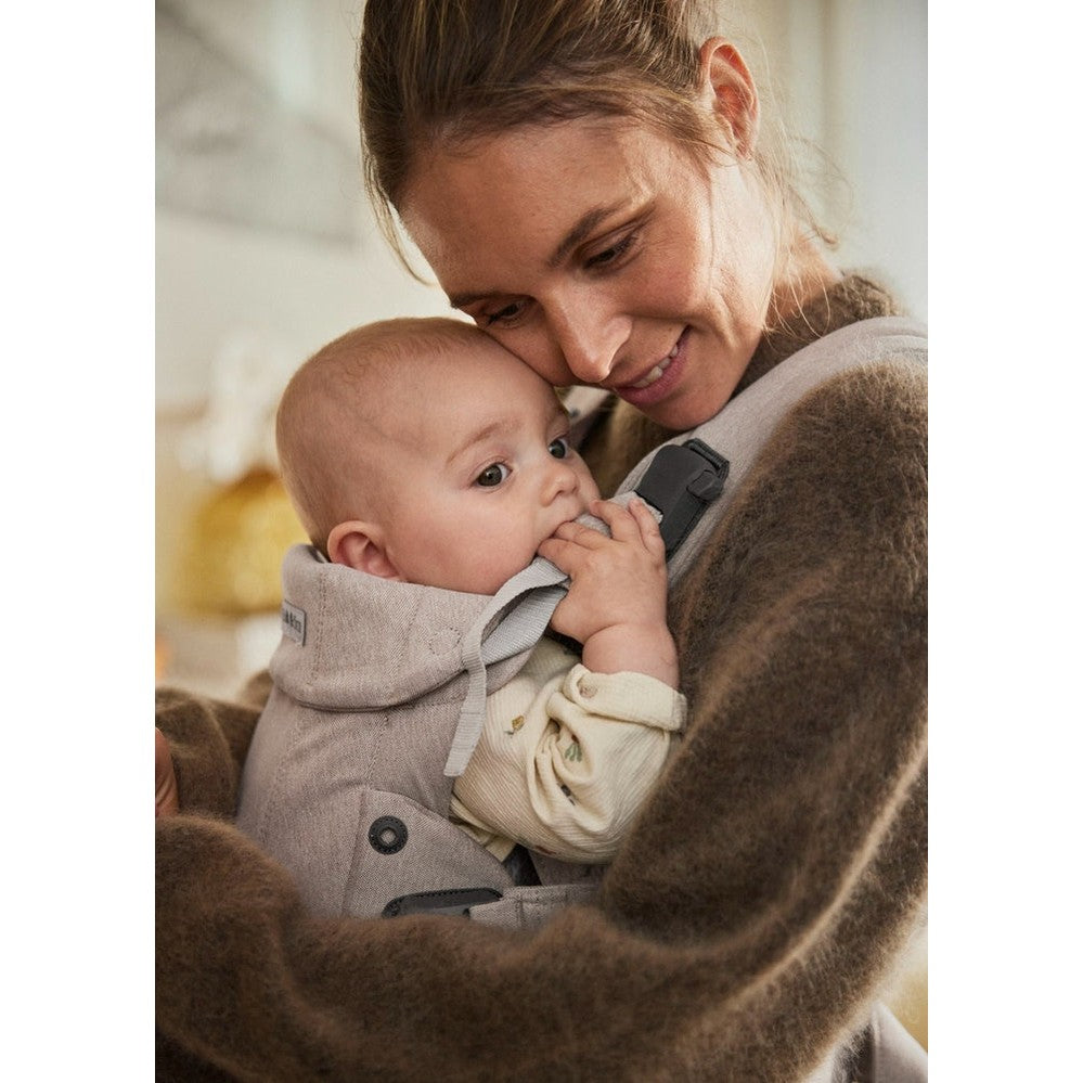 BabyBjörn Harmony baby carrier in gray worn by mother, cradling a smiling infant close against her chest.