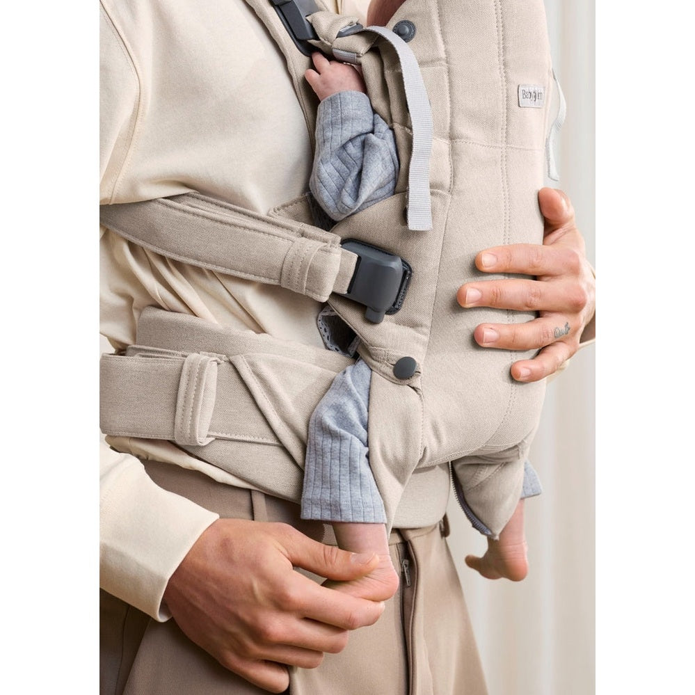 BabyBjörn Baby Carrier Harmony in beige worn by parent with secured waist belt and buckle, baby snug inside.