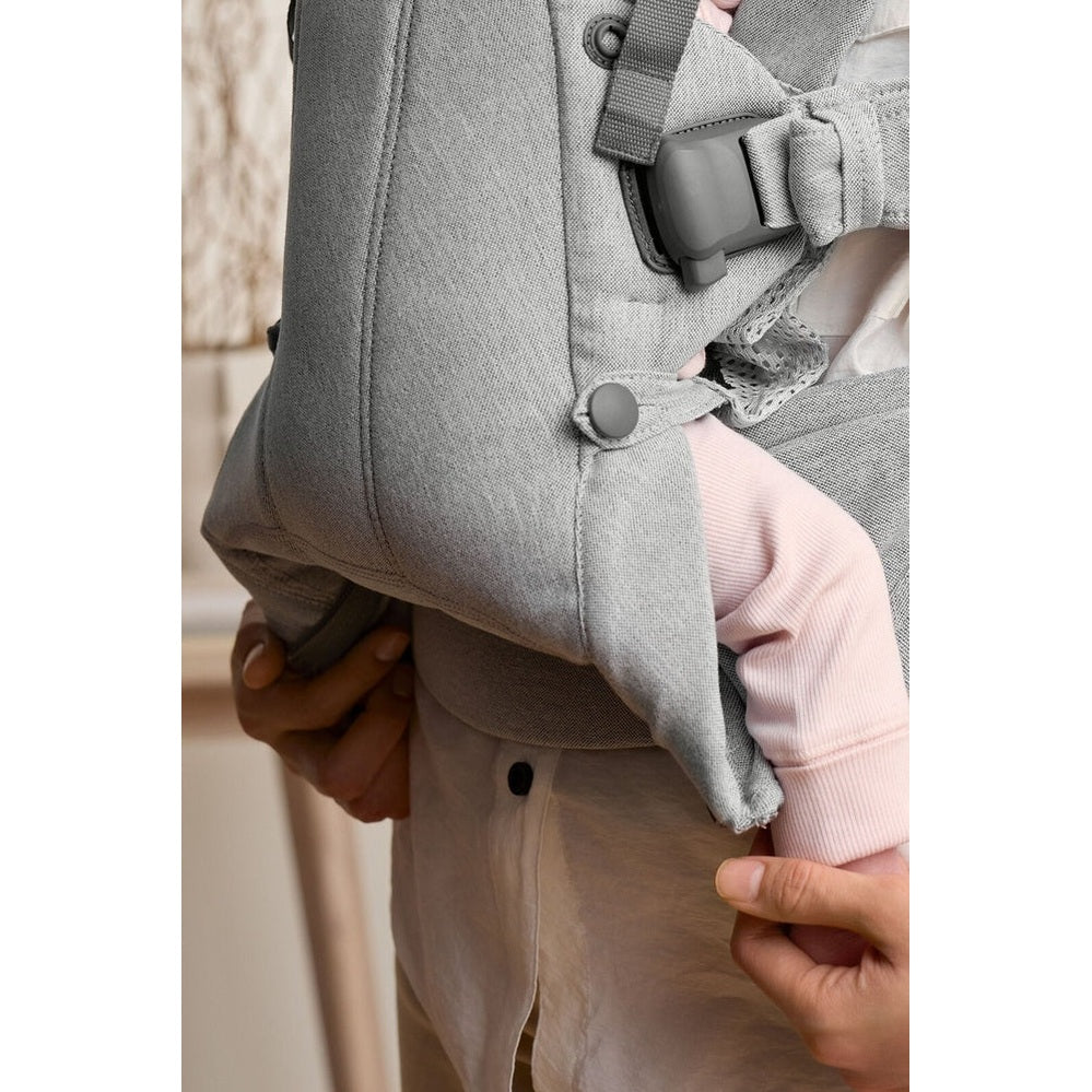 BabyBjörn Baby Carrier Harmony in gray being adjusted by caregiver, securing the leg opening and strap.