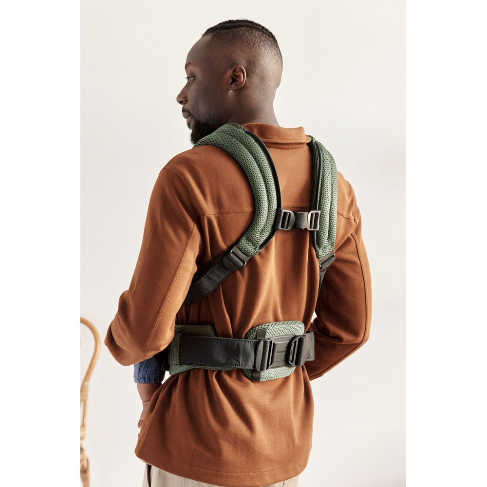 BabyBjörn Baby Carrier Harmony in green with padded straps and hip belt, worn by a man carrying a baby on his back.
