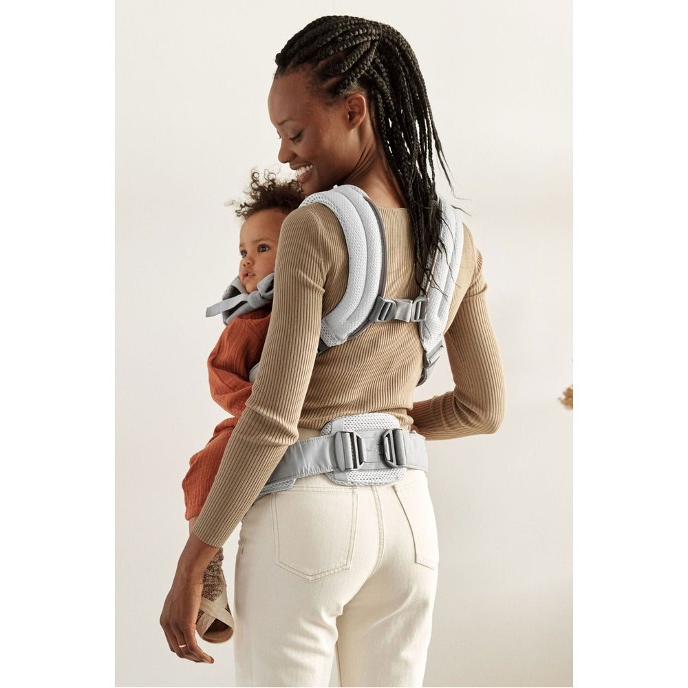 BabyBjörn Harmony baby carrier in light gray worn by smiling mother carrying baby on back with secure straps.