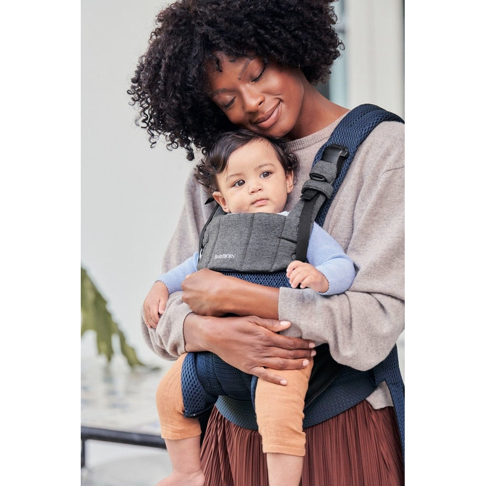 Mother wearing BabyBjörn Baby Carrier Harmony in navy with gray panel, buckled straps, cradling baby securely.