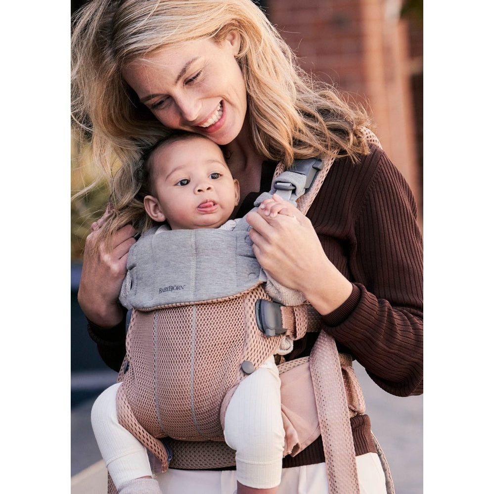 BabyBjörn Baby Carrier Harmony in blush mesh; smiling mom holds baby securely in the buckled front carrier.