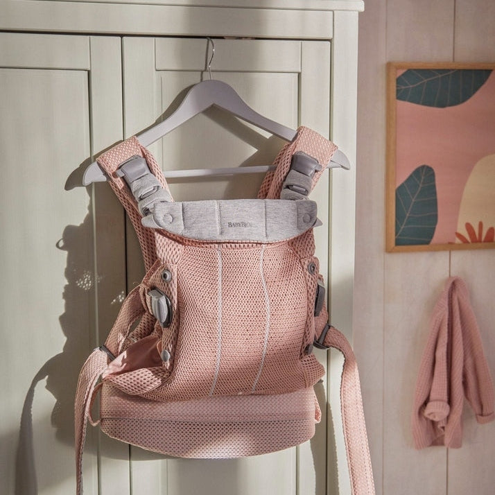BabyBjörn Baby Carrier Harmony in pink mesh hangs on a hanger, showing padded straps and breathable fabric.