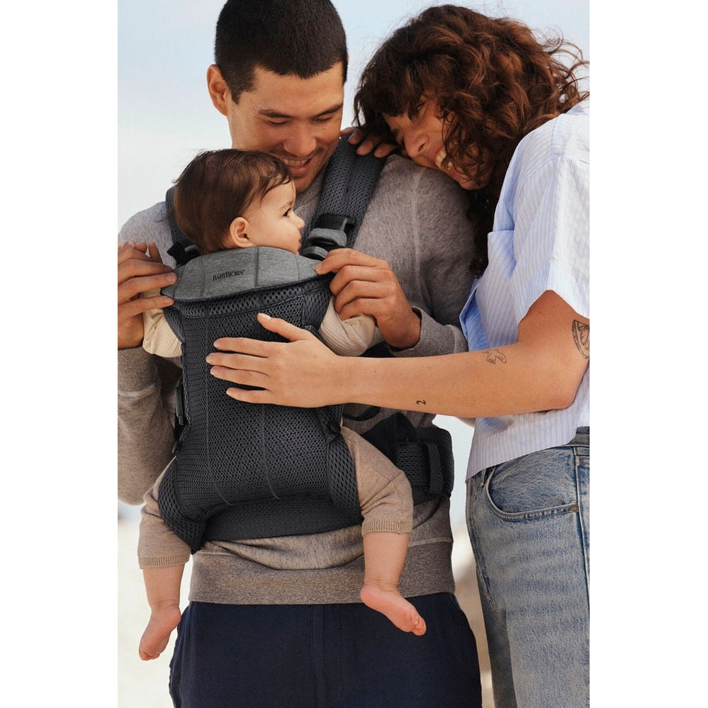 BabyBjörn Harmony baby carrier on beach; parents hold a baby snug against the chest with breathable, ergonomic support.